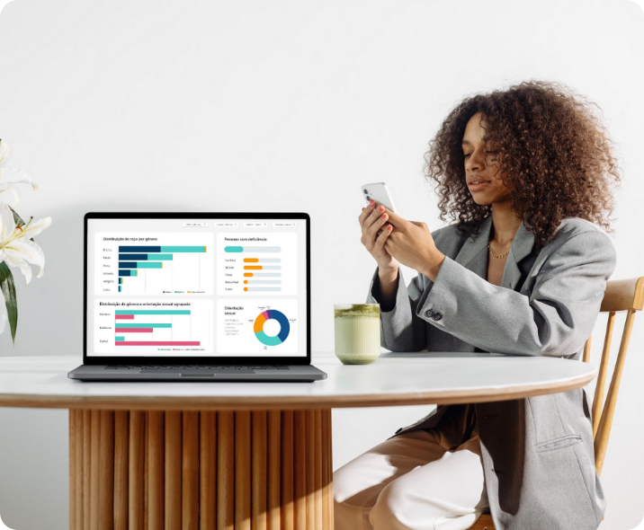 Mulher negra de trajes casuais de negócio, está sentada em uma mesa olhando seu celular. À sua frente há um notebook e na tela está a visão de um dashboard com dados de diversidade