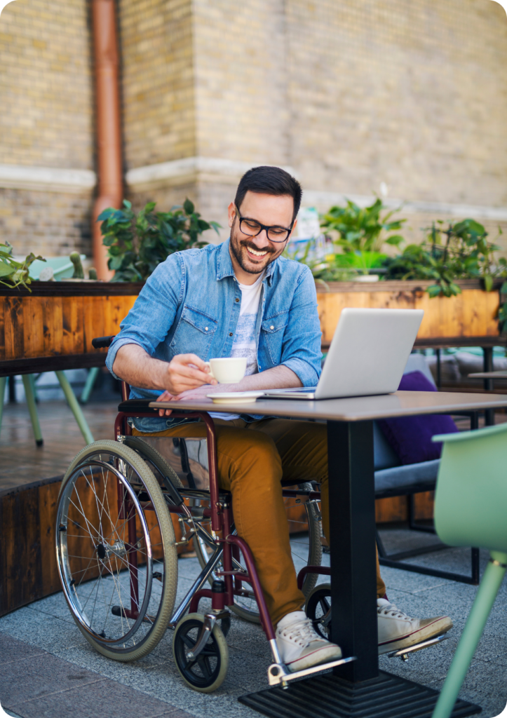 Homem em cadeira de rodas, está em uma mesa de trabalho, em ambiente aberto. A sua frente está um notebook e ele toma um café e sorri.