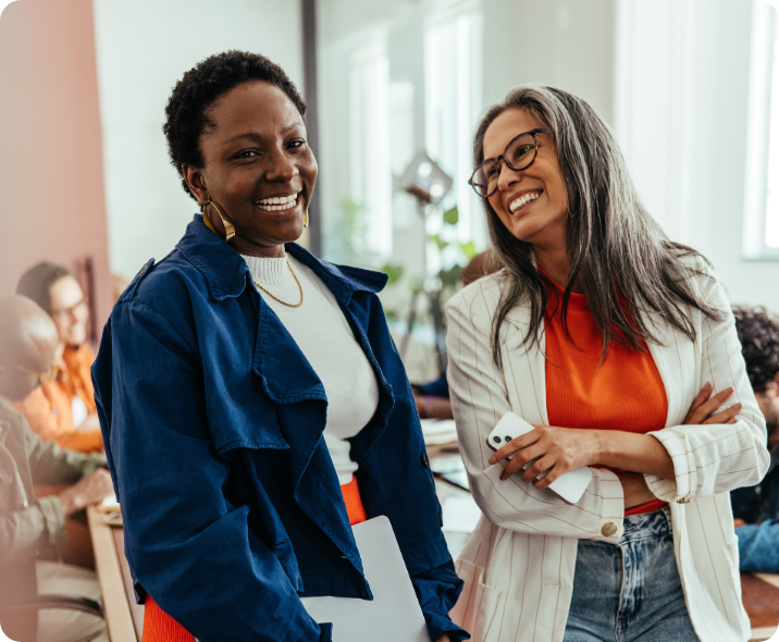 Duas mulheres negras estão sorrindo entre elas. Vestem roupas casuais corporativas e estão em ambiente de escritório.