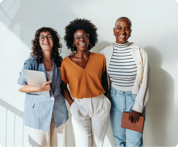 Três mulheres se abraçam de frente para a câmera. As três vestem roupas casuais corporativas e estão sorrindo. Na esquerda há uma mulher branca e ao lado dela, duas mulheres negras.