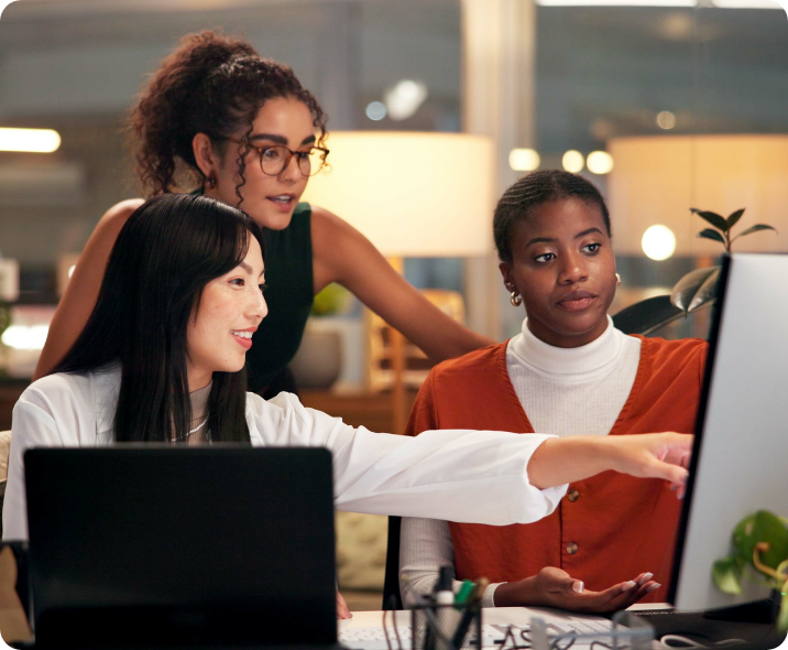 Três mulheres avaliam um conteúdo na tela de um computador. Duas mulheres estão sentadas, uma mulher oriental e uma mulher preta. De pé está a terceira, uma mulher parda. As três estão em ambiente corporativo e vestem roupas casuais corporativas.