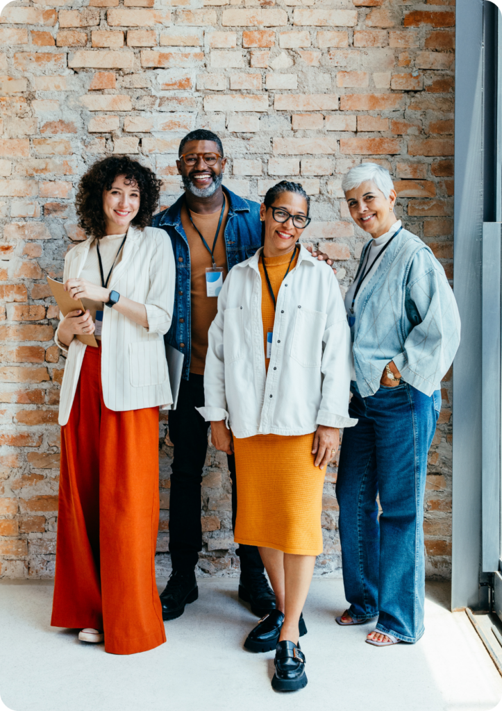 Quatro colegas de trabalho estão lado a lado sorrindo para a câmera. Uma mulher branca, um homem negro mais de 50 anos, uma mulher parda mais de 50 anos e uma mulher branca mais de 50 anos. Todos vestem roupas casuais corporativas.