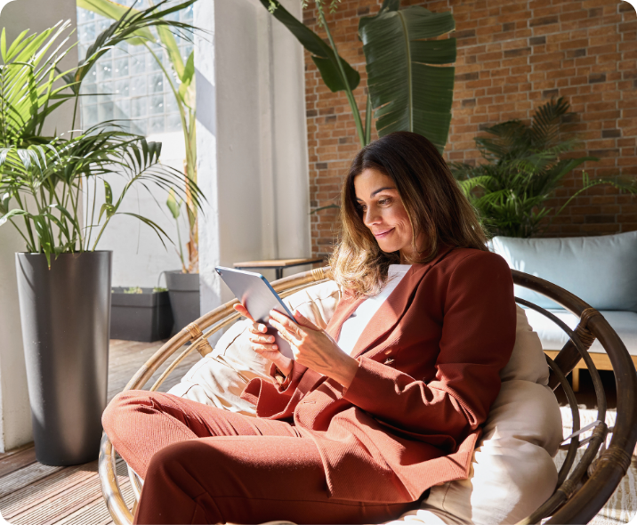 Uma mulher branca está sentava em uma poltrona olhando para um tablet que segura. Sorri enquanto olha o conteúdo do tablet. Veste roupas casuais corporativas.