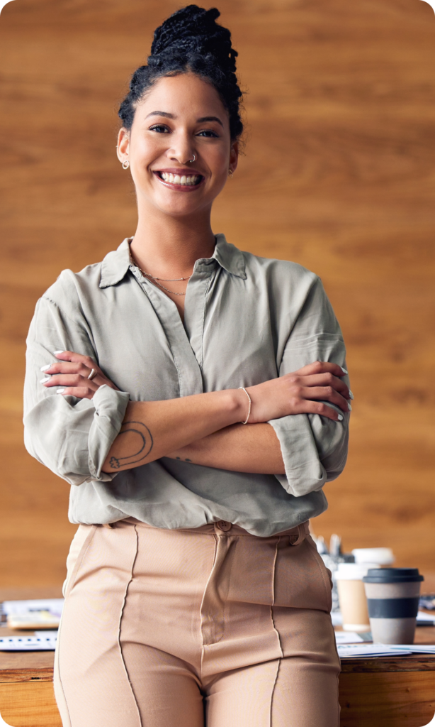 Mulher parda está de pé, encostada em uma mesa. Está sorrindo, de braços cruzados, e veste roupas casuais corporativas.