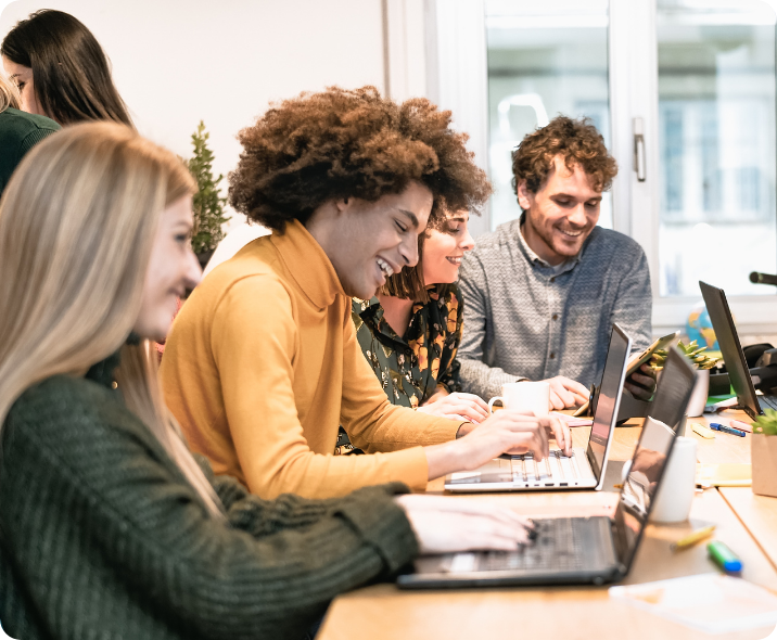 Quatro colegas de trabalho estão com seus notebooks em uma mesa extensa de trabalho. eles sorriem entre eles, vestem roupas casuais corporativas e olham em seus respectivos notebooks.